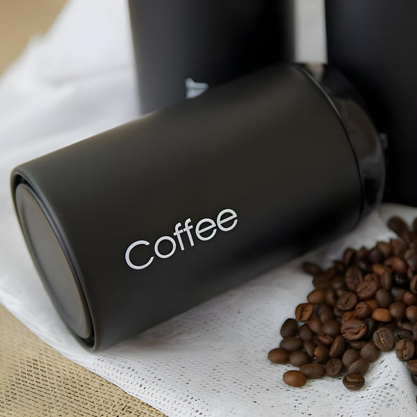 Set of three black metal kitchen canisters with rose gold lids, labelled sugar, tea, and coffee. PALM GIFTS